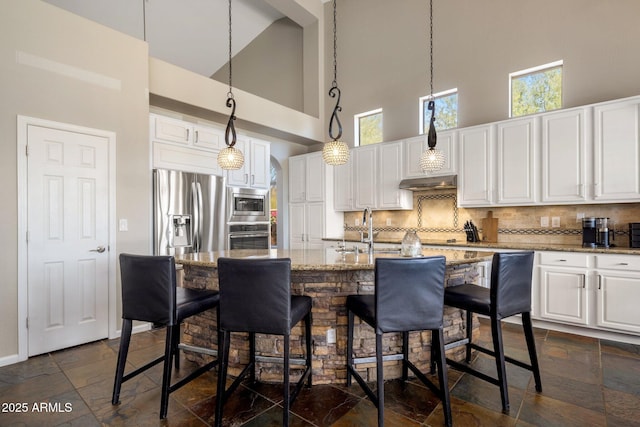 kitchen with under cabinet range hood, stone tile floors, stainless steel appliances, white cabinets, and decorative backsplash
