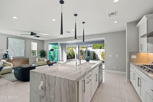 kitchen with sink, a kitchen island with sink, hanging light fixtures, light stone counters, and white cabinets