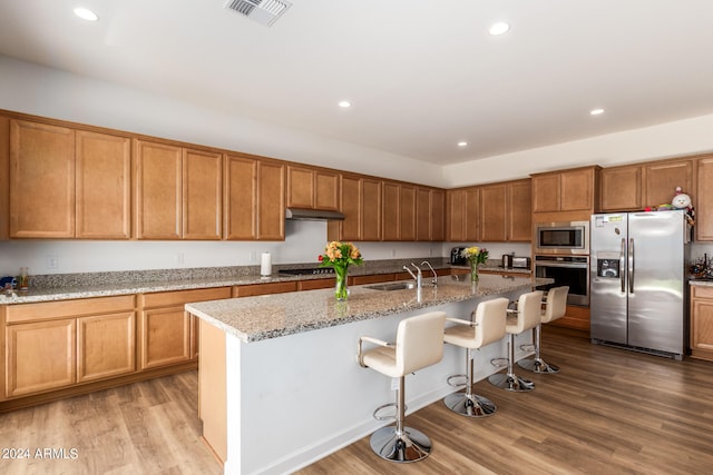 kitchen with light wood-type flooring, stainless steel appliances, light stone counters, a breakfast bar, and a center island with sink