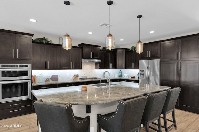 kitchen featuring light wood-type flooring, a kitchen island with sink, decorative light fixtures, and stainless steel appliances