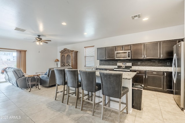 kitchen featuring visible vents, decorative backsplash, appliances with stainless steel finishes, dark brown cabinets, and a kitchen bar