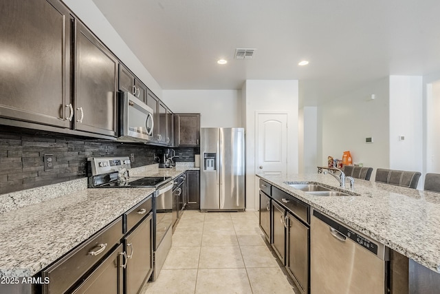 kitchen featuring light tile patterned floors, dark brown cabinetry, a sink, appliances with stainless steel finishes, and backsplash