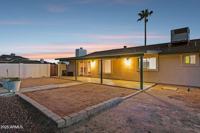 back of house at dusk featuring central AC unit, fence, stucco siding, a chimney, and a patio area