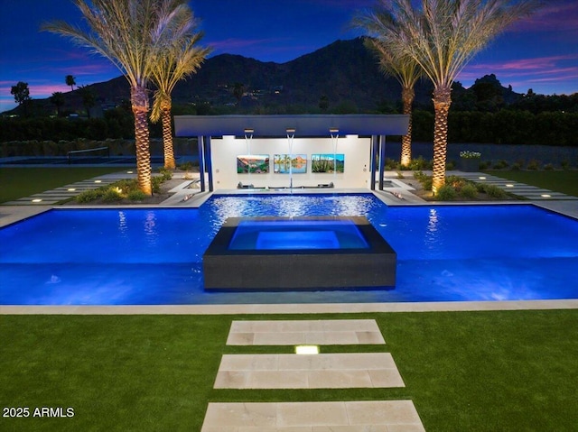 pool at dusk with an in ground hot tub, a mountain view, and a yard