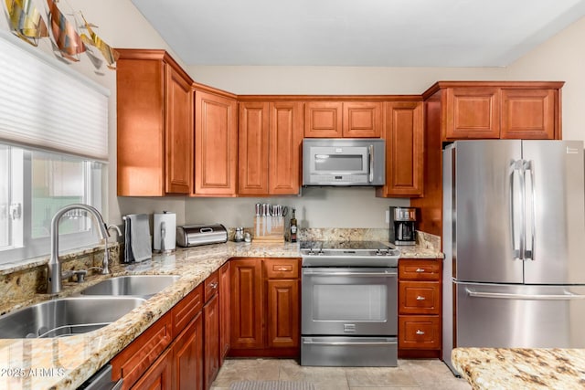 kitchen with stainless steel appliances, light stone counters, brown cabinetry, and a sink