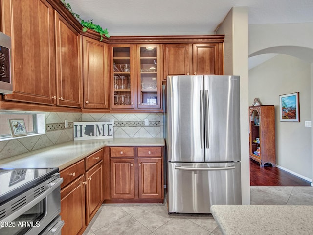 kitchen featuring brown cabinetry, arched walkways, light countertops, tasteful backsplash, and appliances with stainless steel finishes