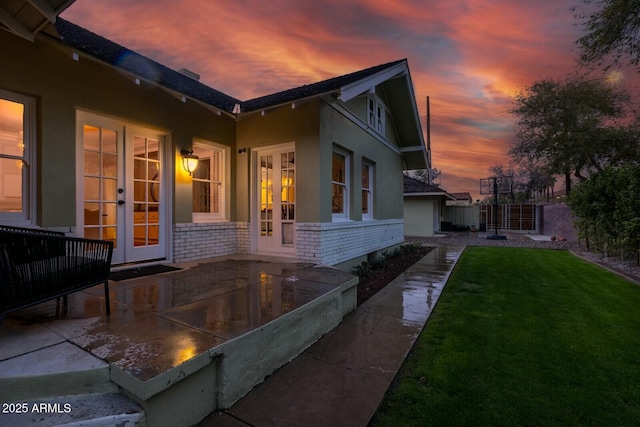 property exterior at dusk with fence, french doors, a lawn, and stucco siding