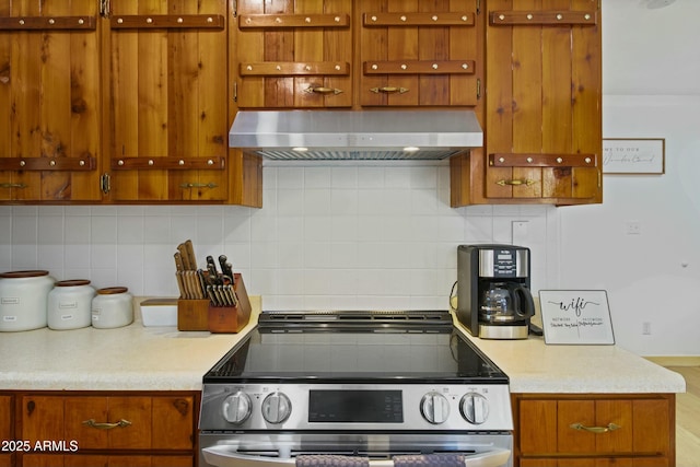 kitchen featuring wall chimney range hood, stainless steel range with electric cooktop, light countertops, brown cabinetry, and backsplash