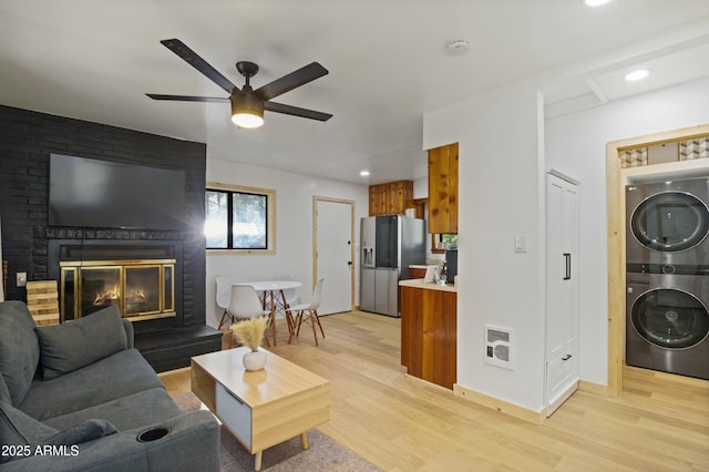 living room featuring a ceiling fan, stacked washer and dryer, a brick fireplace, baseboards, and light wood-style flooring