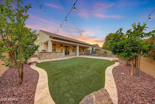 back of house at dusk featuring a tiled roof, a lawn, a fenced backyard, a patio, and stucco siding