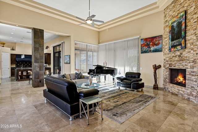 living area with a stone fireplace, a high ceiling, a ceiling fan, a tray ceiling, and decorative columns