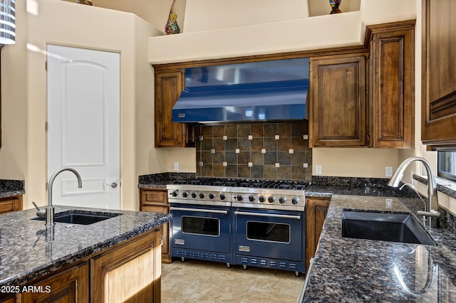 kitchen with dark stone counters, wall chimney range hood, range with two ovens, and a sink