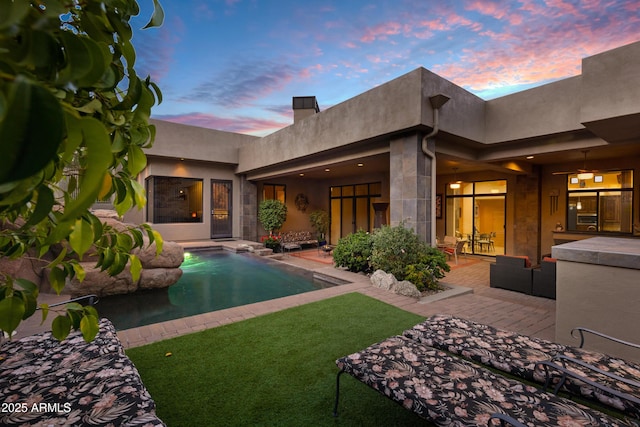 back of house at dusk with stucco siding, an outdoor pool, a lawn, and a patio