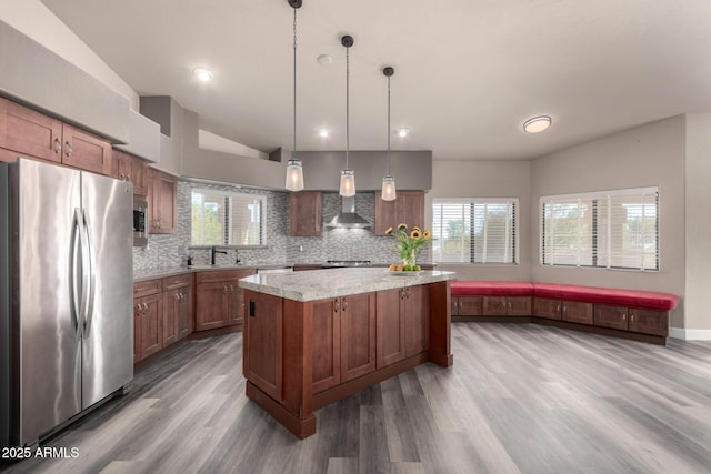 kitchen with a center island, decorative light fixtures, stainless steel appliances, brown cabinetry, and wall chimney range hood