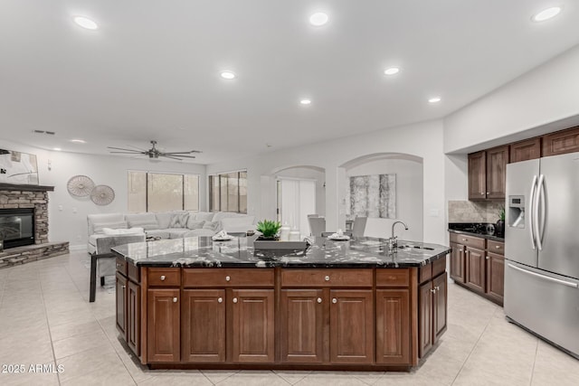 kitchen with stainless steel fridge, dark stone counters, an island with sink, a stone fireplace, and a sink