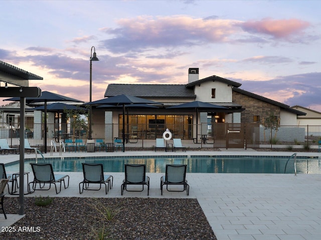 pool at dusk featuring a patio area, fence, and a community pool