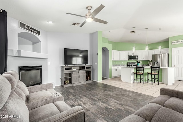 living room with visible vents, a ceiling fan, a glass covered fireplace, vaulted ceiling, and baseboards