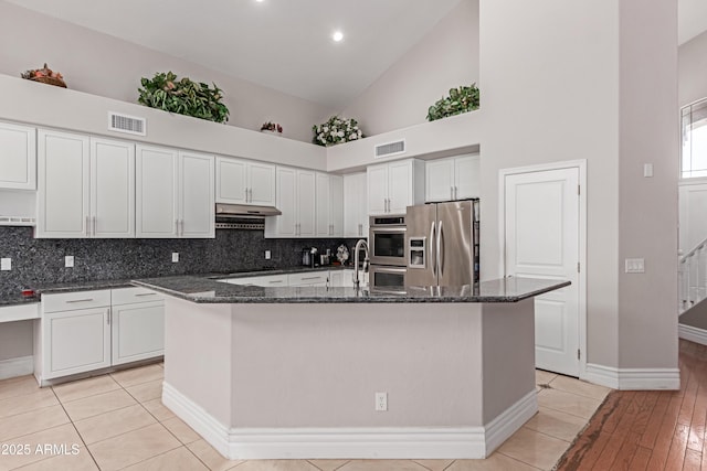 kitchen featuring a center island with sink, under cabinet range hood, dark stone countertops, stainless steel appliances, and visible vents
