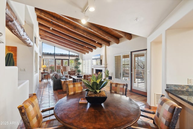 tiled dining space with a warm lit fireplace, lofted ceiling with beams, visible vents, and track lighting