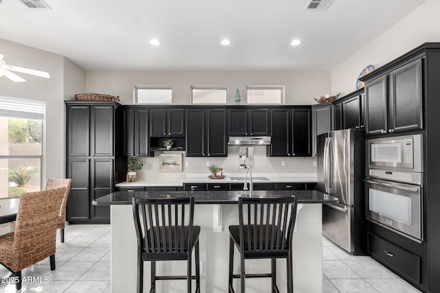 kitchen with light tile patterned floors, a breakfast bar, appliances with stainless steel finishes, dark stone countertops, and a center island with sink