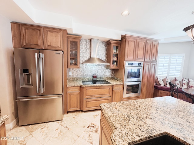 kitchen featuring appliances with stainless steel finishes, wall chimney range hood, light stone countertops, decorative backsplash, and glass insert cabinets