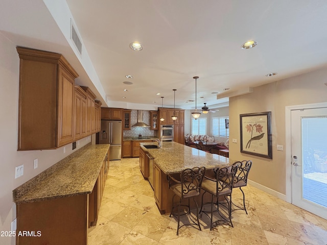 kitchen featuring appliances with stainless steel finishes, wall chimney range hood, a sink, brown cabinets, and ceiling fan