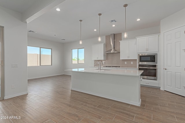 kitchen featuring wall chimney range hood, appliances with stainless steel finishes, pendant lighting, white cabinets, and a center island with sink