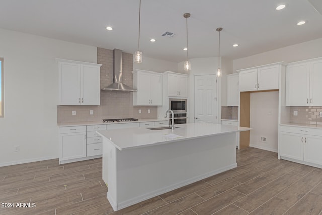 kitchen with wall chimney range hood, an island with sink, sink, decorative light fixtures, and white cabinets