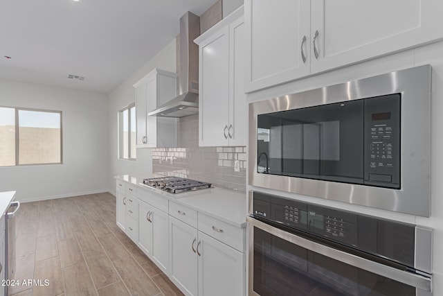 kitchen with wall chimney exhaust hood, light hardwood / wood-style flooring, stainless steel appliances, white cabinets, and tasteful backsplash