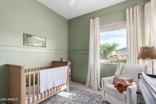 bedroom with a crib, a ceiling fan, wainscoting, and carpet flooring