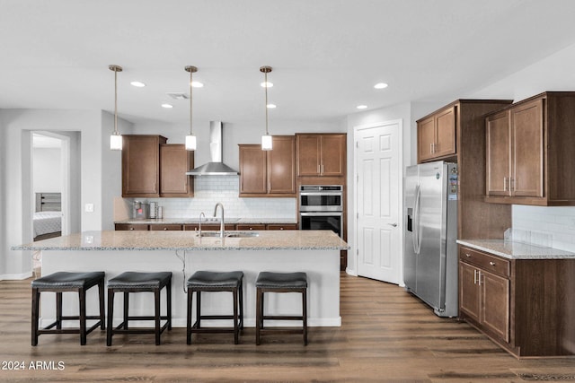 kitchen with hanging light fixtures, appliances with stainless steel finishes, wall chimney exhaust hood, and a kitchen island with sink