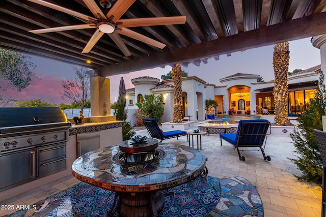 patio terrace at dusk with an outdoor kitchen, an outdoor bar, ceiling fan, and a fire pit