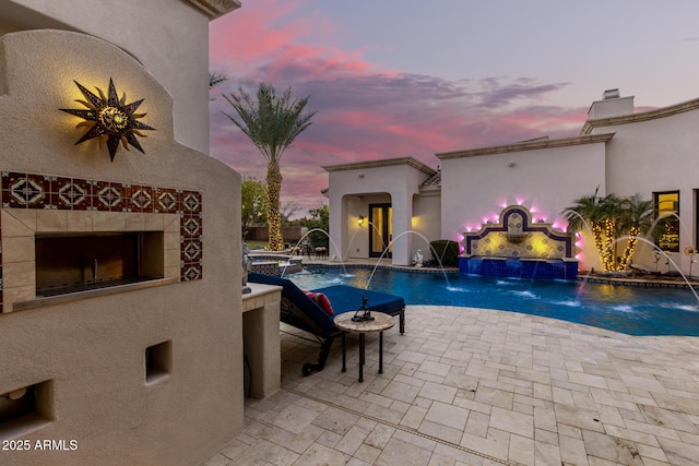 pool at dusk featuring a patio, pool water feature, and a fireplace