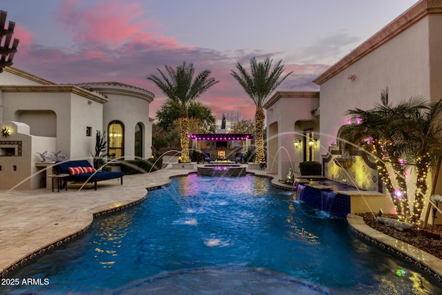 pool at dusk featuring exterior fireplace, a patio area, pool water feature, and an in ground hot tub