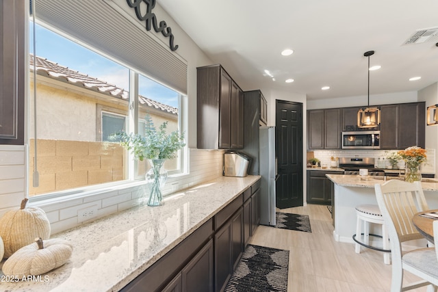 kitchen with light stone countertops, appliances with stainless steel finishes, tasteful backsplash, dark brown cabinets, and hanging light fixtures