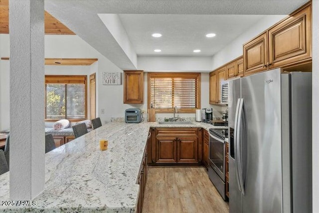 kitchen with kitchen peninsula, sink, light wood-type flooring, light stone countertops, and appliances with stainless steel finishes