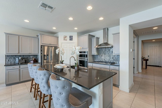 kitchen featuring an island with sink, appliances with stainless steel finishes, gray cabinetry, and wall chimney range hood