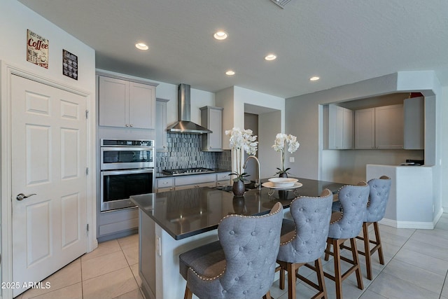 kitchen featuring gray cabinets, an island with sink, and wall chimney exhaust hood