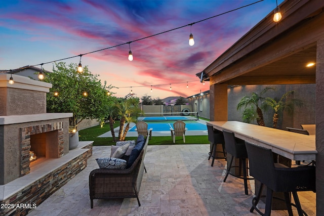 patio terrace at dusk featuring a tennis court, fence, a warm lit fireplace, and outdoor dry bar