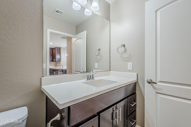 half bathroom with vanity, toilet, a textured wall, and visible vents