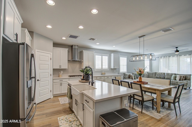 kitchen with a center island with sink, appliances with stainless steel finishes, hanging light fixtures, white cabinets, and wall chimney range hood