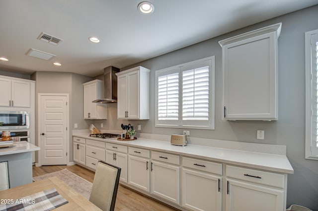 kitchen with appliances with stainless steel finishes, white cabinets, light wood-type flooring, and wall chimney exhaust hood