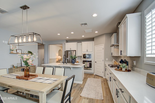 kitchen featuring appliances with stainless steel finishes, an island with sink, light wood-type flooring, pendant lighting, and white cabinets