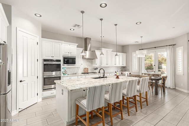 kitchen with wall chimney exhaust hood, an island with sink, pendant lighting, stainless steel appliances, and light stone countertops