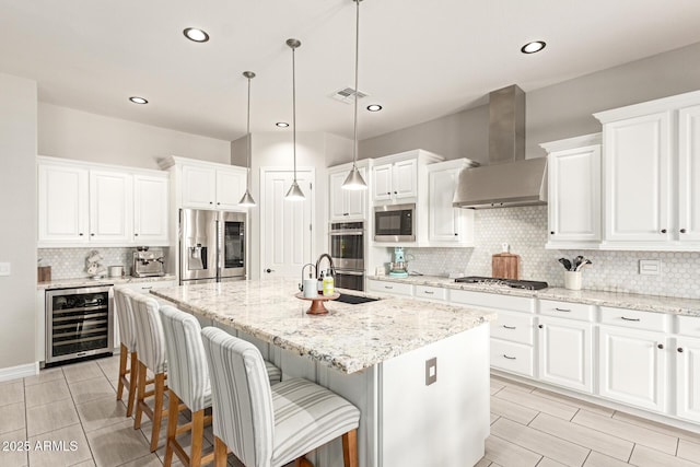 kitchen featuring wine cooler, decorative light fixtures, stainless steel appliances, a kitchen island with sink, and wall chimney range hood