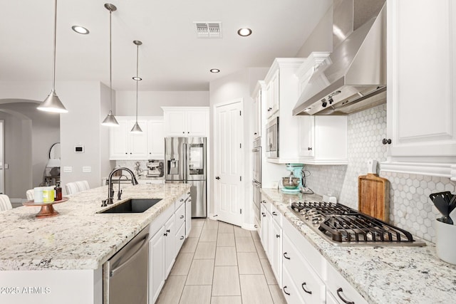 kitchen featuring wall chimney range hood, appliances with stainless steel finishes, hanging light fixtures, white cabinets, and a center island with sink