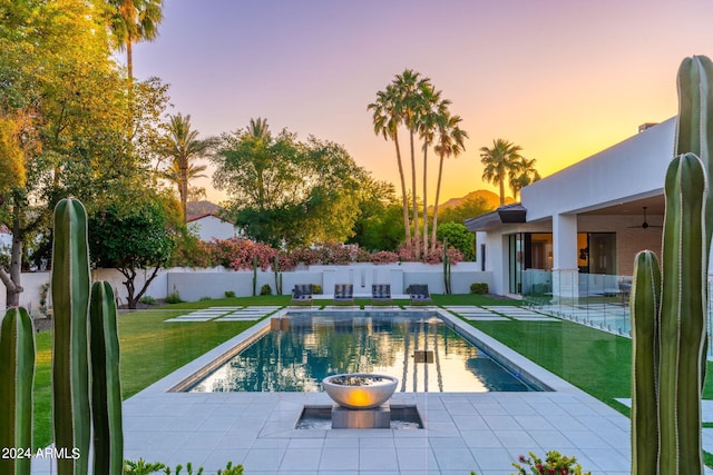 pool at dusk with a yard, ceiling fan, and a patio