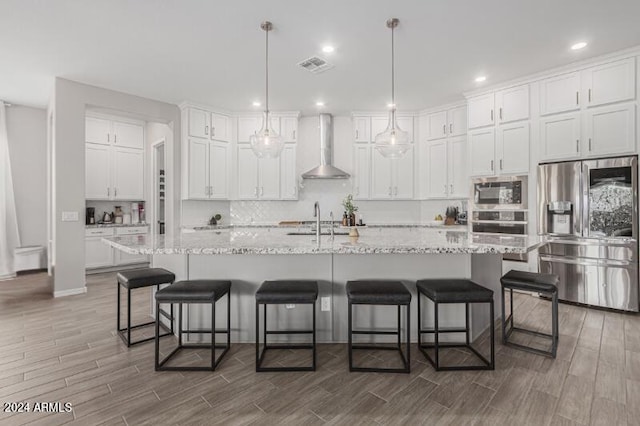 kitchen featuring a center island with sink, white cabinets, wall chimney range hood, and appliances with stainless steel finishes