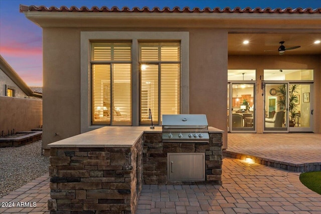 patio terrace at dusk with ceiling fan, a grill, and exterior kitchen