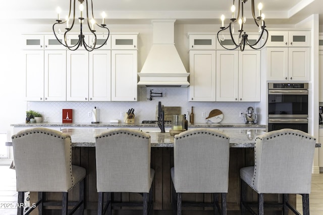 kitchen featuring a kitchen island with sink, a notable chandelier, premium range hood, and stainless steel double oven